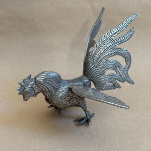 Könnte beinhalten: Eine detaillierte silberfarbene Hahnfigur. Der Hahn hat einen strukturierten Körper, einen markanten Kamm und einen großen, gefächerten Schwanz. Die Flügel sind ausgebreitet und der Vogel scheint sich zu bewegen. Die Figur ist ein dekorativer Gegenstand.