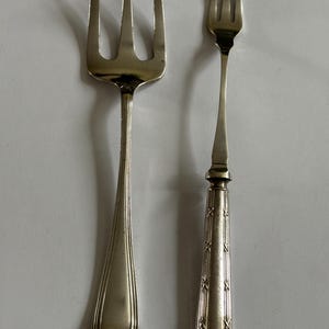 Puede incluir: Dos tenedores de metal plateado colocados uno al lado del otro. El tenedor de la izquierda es una pieza grande de tres dientes con un mango liso y ensanchado. El de la derecha es más pequeño, con tres dientes y un mango con patrones de cruz grabados.