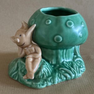 Puede incluir: Una caprichosa figura de cerámica que representa un hongo verde con una pequeña abertura en la parte superior. Una figura de elfo de color beige está sentada sobre una base verde, apoyada contra el hongo. La figura tiene un acabado brillante.