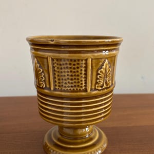 May include: A brown ceramic goblet with a pedestal base. The goblet features embossed leaf and geometric designs. Horizontal ridges encircle the base and the top rim. The goblet is displayed on a wooden surface.