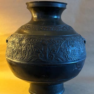 May include: A dark bronze vase with a rounded body and flared neck. The vase has detailed relief carvings of mythical creatures and patterns. It stands on a pedestal base and has small handles. The vase is a dark bronze colour.