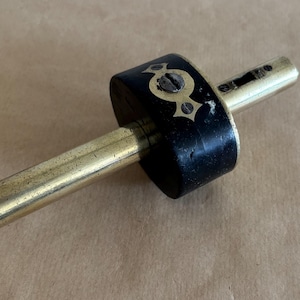May include: A marking gauge tool featuring a cylindrical brass rod passing through a circular black wooden block. The block is secured with a decorative brass plate and a single screw, while the brass rod shows visible wear and metallic texture.