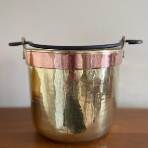 May include: A hammered brass bucket with a copper band around the top. The bucket has a black handle and is on a wooden surface. The bucket is likely for decorative use.