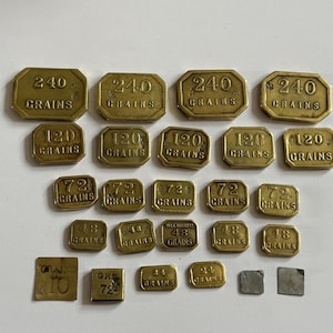 May include: A collection of rectangular brass weights of varying sizes, each stamped with its specific weight in grains, including values of 240, 120, 72, 48, 24, 10, and 5 grains, alongside two small square silver-toned metal pieces.