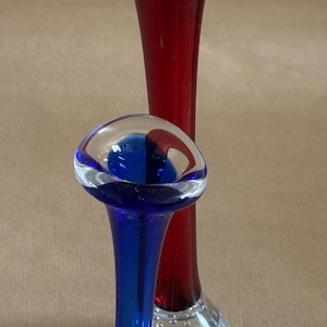 Op de afbeelding: Twee glazen vaasjes met uitlopende randen en heldere, met luchtbellen getextureerde voeten. De langere vaas is dieprood, terwijl de kortere vaas felblauw is. Een ronde sticker met de tekst SWEDISH ART GLASS is zichtbaar op de voet.