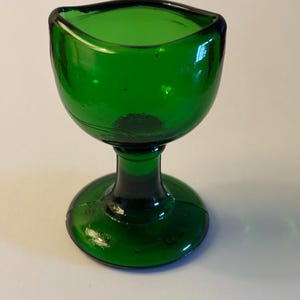 Op de afbeelding: Een kleine, levendig smaragdgroene glazen oogbad met een afgeronde kom en een voorgevormde rand die is ontworpen om op de oogkas te passen, ondersteund door een korte, dikke steel en een brede, cirkelvormige voet van glad glas.