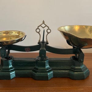 May include: A vintage-style balance scale featuring a dark green cast iron base and central support column. Two polished brass bowls are suspended from the horizontal beam, which includes a central vertical pointer and decorative brass finial.