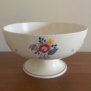 May include: A white ceramic pedestal bowl with a floral design in shades of pink, purple, yellow, and green. The bowl has a scalloped edge and a purple trim.
