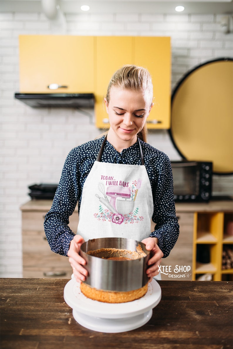 May include: A white apron with the text "Today I will bake" and a pink mixer design. The apron is worn by a person in a kitchen, holding a cake with a metal ring. The kitchen has yellow cabinets and a black oven.