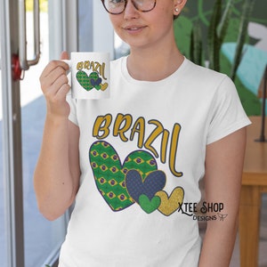 以下が含まれることがあります： 「BRAZIL」の文字が金と青で描かれ、緑、青、金色のハートのグラフィックが描かれた白いTシャツ。お揃いのマグカップが掲げられています。デザインはブラジルの国旗を特徴としています。