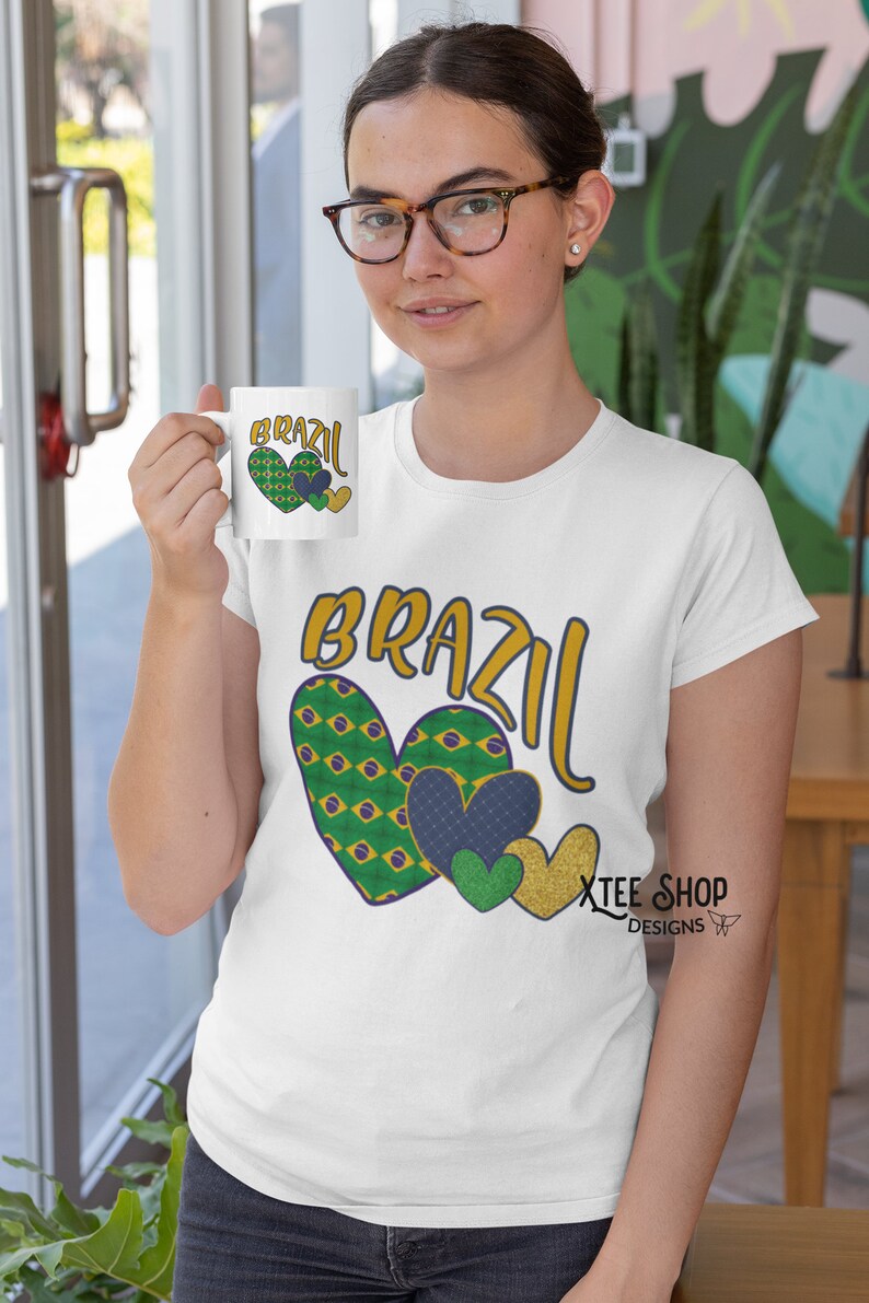 以下が含まれることがあります： 「BRAZIL」の文字が金と青で描かれ、緑、青、金色のハートのグラフィックが描かれた白いTシャツ。お揃いのマグカップが掲げられています。デザインはブラジルの国旗を特徴としています。