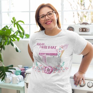 May include: A light gray t-shirt with the text "TODAY... I WILL BAKE" above a watercolor illustration of a pink stand mixer, whisk, and spatula. The design is surrounded by a floral wreath. The person is wearing glasses.