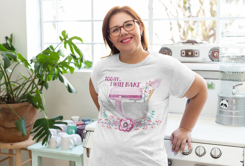 May include: A light gray t-shirt with the text "TODAY... I WILL BAKE" above a watercolor illustration of a pink stand mixer, whisk, and spatula. The design is surrounded by a floral wreath. The person is wearing glasses.
