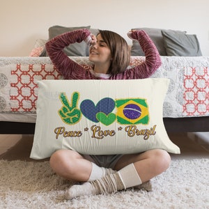 以下が含まれることがあります： 「Peace Love Brazil」の文字とピースサイン、2つのハート、ブラジル国旗が描かれた長方形の枕。 ピースサインは緑と金、ハートは青と緑、国旗は黄色、緑、青、白です。