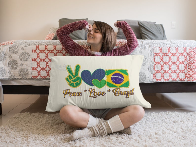 以下が含まれることがあります： 「Peace Love Brazil」の文字とピースサイン、2つのハート、ブラジル国旗が描かれた長方形の枕。 ピースサインは緑と金、ハートは青と緑、国旗は黄色、緑、青、白です。