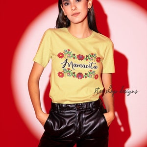 May include: A yellow t-shirt with a floral design and the text "Mamacita" in black script. The shirt is being worn by a person with long dark hair and is paired with black leather pants.