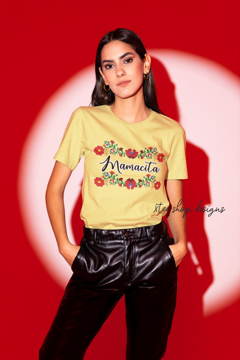 May include: A yellow t-shirt with a floral design and the text "Mamacita" in black script. The shirt is being worn by a person with long dark hair and is paired with black leather pants.