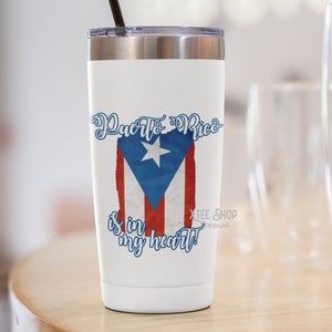May include: White travel mug with a silver lid and a black straw. The mug features the Puerto Rican flag design with the text "Puerto Rico is in my heart!" in blue script. The mug is on a wooden surface.