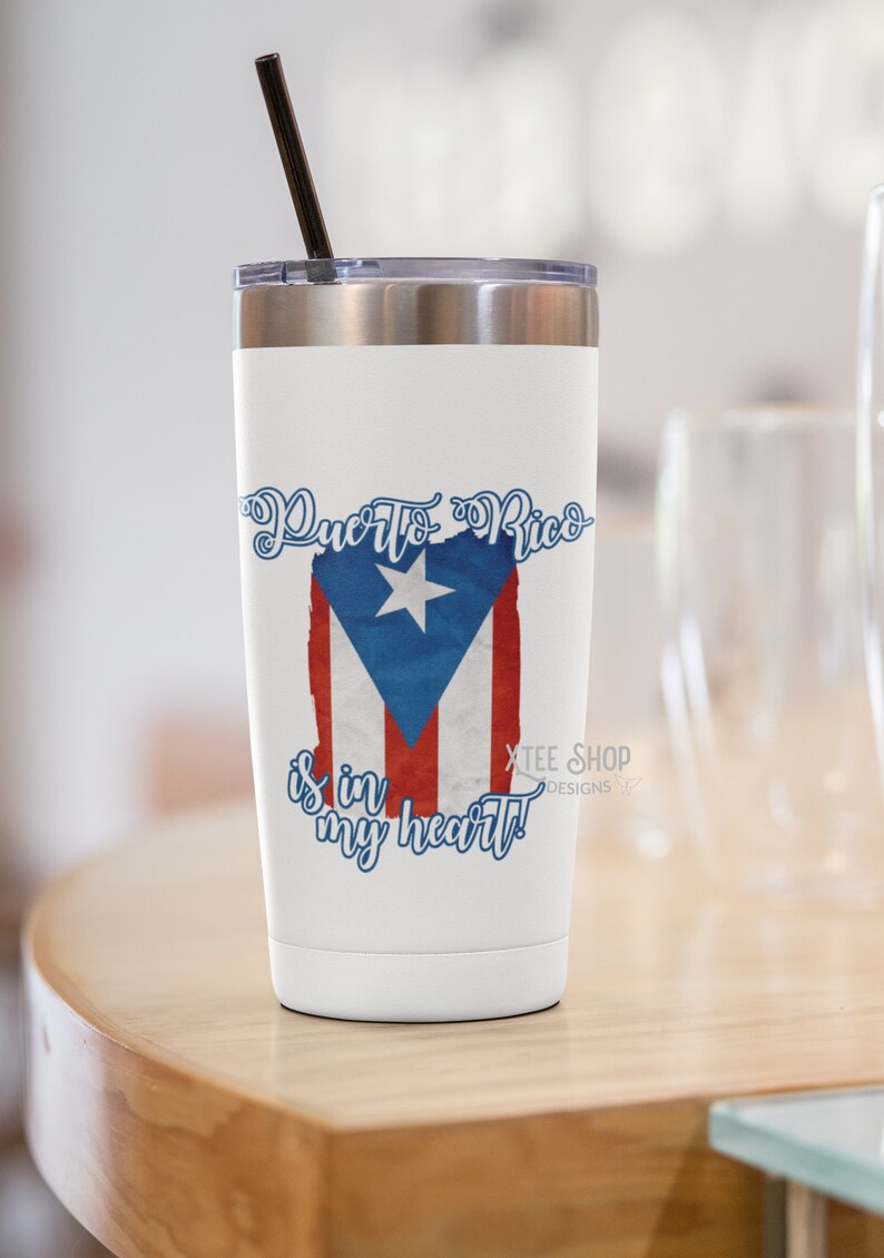 May include: White travel mug with a silver lid and a black straw. The mug features the Puerto Rican flag design with the text "Puerto Rico is in my heart!" in blue script. The mug is on a wooden surface.
