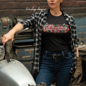 May include: A woman wearing a black t-shirt with the text "Chingona" in colorful embroidery, a black and white plaid shirt, blue jeans, and a welding mask. She is standing next to a silver motorcycle.