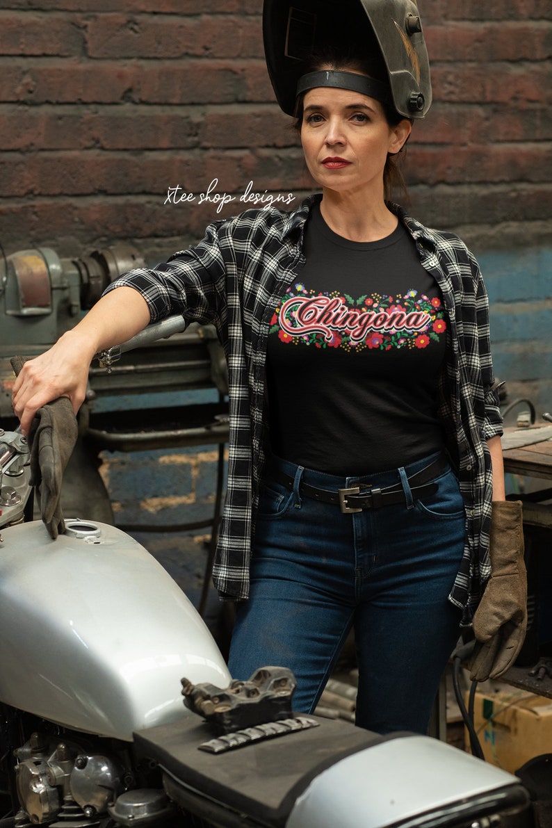 May include: A woman wearing a black t-shirt with the text "Chingona" in colorful embroidery, a black and white plaid shirt, blue jeans, and a welding mask. She is standing next to a silver motorcycle.