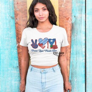 May include: White t-shirt with a graphic design featuring the words "Peace Love Puerto Rico" and a peace sign, heart, and Puerto Rican flag. The shirt is worn by a person in light blue denim shorts.