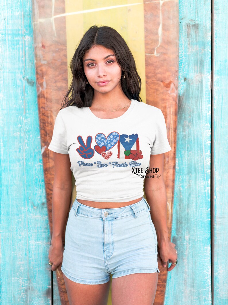 May include: White t-shirt with a graphic design featuring the words "Peace Love Puerto Rico" and a peace sign, heart, and Puerto Rican flag. The shirt is worn by a person in light blue denim shorts.