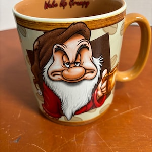 May include: A ceramic coffee mug featuring Grumpy from Snow White. The mug has a brown handle and interior, with a beige exterior decorated with coffee beans. The words "Wake Up Grumpy" are printed on the rim. The Grumpy character is depicted in his signature grumpy expression.