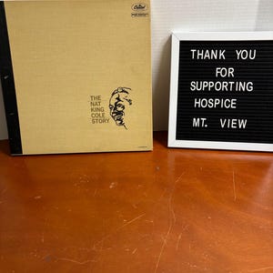 以下が含まれることがあります： タン色の表紙と黒い背表紙が付いた、The Nat King Cole Storyというタイトルのヴィンテージビニールレコードアルバム。アルバムの表紙には、ナット・キング・コールの肖像画が描かれています。白い文字で「THANK YOU FOR SUPPORTING HOSPICE MT. VIEW」と書かれた黒いレターボード。