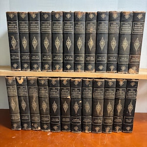 May include: A collection of antique books, featuring dark covers with gold lettering and decorative patterns. The books are arranged on a wooden shelf, showcasing the spines with titles like "The Ridpath Library of Universal Literature."