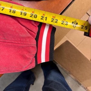 Puede incluir: Una manga de chaqueta roja con un pu&ntilde;o a rayas negras, blancas y rojas se mide con una cinta m&eacute;trica amarilla. La manga mide aproximadamente 56 cm de largo.