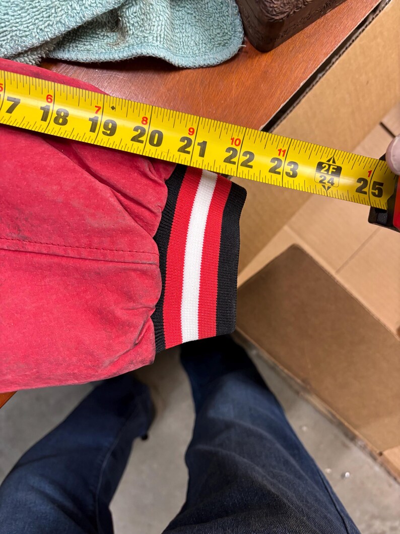 Puede incluir: Una manga de chaqueta roja con un pu&ntilde;o a rayas negras, blancas y rojas se mide con una cinta m&eacute;trica amarilla. La manga mide aproximadamente 56 cm de largo.