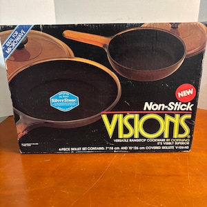May include: A box set of Visions non-stick skillets. The box features images of the cookware, with the words "Non-Stick" and "VISIONS" prominently displayed. The set includes 7-inch and 10-inch skillets, ideal for microwave use.