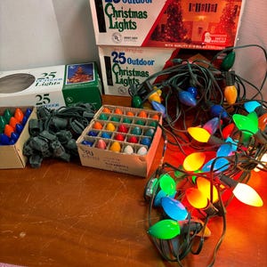 May include: Assorted Christmas light sets with colorful bulbs and green wiring. Boxes of 25 outdoor lights are stacked, featuring images of a house and Christmas tree. A measuring tape is visible at the bottom.