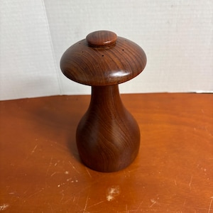 Może przedstawiać: Drewniana solniczka lub pieprzniczka w kształcie grzyba. Shaker wykonany jest z ciemnobrązowego drewna o gładkim, polerowanym wykończeniu. Na górze znajduje się małe, okrągłe pokrętło i otwory do dozowania. Podstawa ma zaokrąglony kształt.