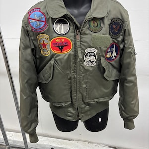 May include: An olive green bomber jacket with various patches. Patches feature text and images of aircraft, a star, and the Eiffel Tower. The jacket has a zip closure, two front pockets, and ribbed cuffs and hem.