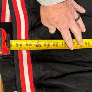 Puede incluir: Chaqueta negra con ribete a rayas rojas y blancas. Una cinta m&eacute;trica amarilla muestra una longitud de aproximadamente 53 cm. Una persona est&aacute; midiendo la chaqueta.