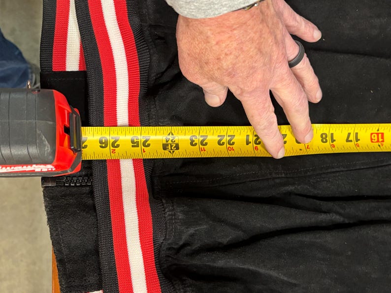 Puede incluir: Chaqueta negra con ribete a rayas rojas y blancas. Una cinta m&eacute;trica amarilla muestra una longitud de aproximadamente 53 cm. Una persona est&aacute; midiendo la chaqueta.