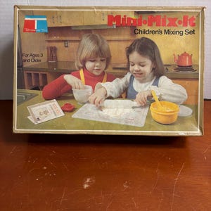 以下が含まれることがあります： TupperwareのMini-Mix-It子供用ミキシングセットのヴィンテージボックス。ボックスには、セットで遊んでいる2人の少女のイラストが描かれています。ボックスには「Mini-Mix-It Children's Mixing Set」と「For Ages 3 and Older」の文字があります。
