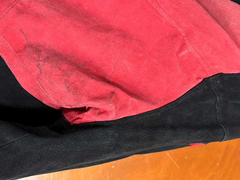 Puede incluir: Primer plano de una chaqueta negra con forro rojo. El forro rojo es visible, mostrando una superficie texturizada con manchas m&aacute;s oscuras. La chaqueta parece ser de ante o un material similar.