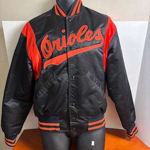 Può includere: Giacca da baseball nera in raso con dettagli arancioni e la scritta "Orioles" in arancione sul petto. La giacca ha una chiusura con bottoni, polsini a righe e un colletto con strisce arancioni e blu navy.