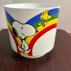 Może przedstawiać: Biała cylindryczna doniczka z kolorową grafiką przedstawiającą Snoopy'ego i Woodstocka. Wzór zawiera żywą tęczę za postaciami, z niebieskimi, zielonymi, żółtymi i czerwonymi pasami owijającymi się wokół zewnętrznej powierzchni przedmiotu.