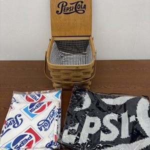 Op de afbeelding: Een houten Pepsi-Cola picknickmand met een gingham voering, een Pepsi-Cola deken en een Pepsi-Cola shirt met patroon. De mand heeft een scharnierend deksel en een handvat. Het shirt en de deken zitten in plastic verpakking.
