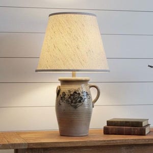May include: A beige ceramic lamp with a floral design and a tan lampshade. The lamp sits on a wooden table with two books. The lamp has a handle and a grey trim on the lampshade.