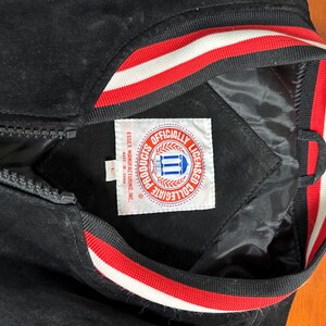 Puede incluir: Chaqueta universitaria negra con cierre de cremallera. El cuello y los pu&ntilde;os tienen rayas rojas y blancas. La chaqueta tiene una etiqueta que dice "Officially Licensed Collegiate Products".