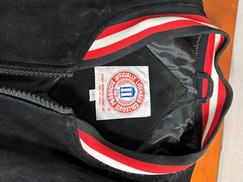 Puede incluir: Chaqueta universitaria negra con cierre de cremallera. El cuello y los pu&ntilde;os tienen rayas rojas y blancas. La chaqueta tiene una etiqueta que dice "Officially Licensed Collegiate Products".