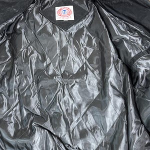 Puede incluir: Interior de chaqueta negra con forro acolchado. La chaqueta tiene un cuello a rayas rojas y blancas. Una etiqueta est&aacute; cosida en la chaqueta con el texto "M" dentro de un cuadrado.