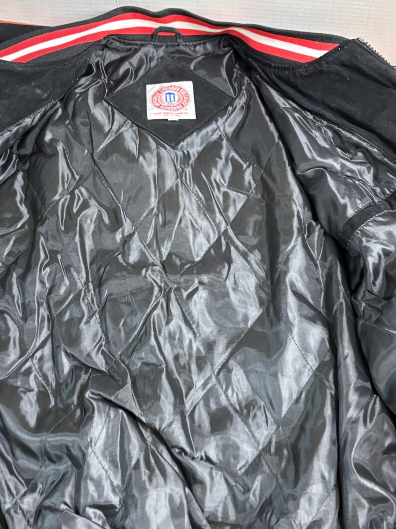 Puede incluir: Interior de chaqueta negra con forro acolchado. La chaqueta tiene un cuello a rayas rojas y blancas. Una etiqueta est&aacute; cosida en la chaqueta con el texto "M" dentro de un cuadrado.