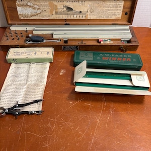 Può includere: Un set di lettering Leroy vintage in una custodia di legno, con vari strumenti, tra cui un accessorio per penna a compasso, matite e un righello. Include anche un astuccio per matite in metallo verde con l'etichetta "A.W. Faber Winner".