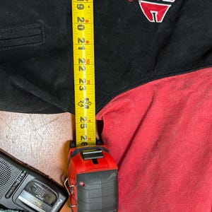 Puede incluir: Chaqueta negra y roja con un logotipo en la parte superior izquierda del pecho. Una cinta m&eacute;trica amarilla muestra una longitud de aproximadamente 63,5 cm. Un peque&ntilde;o reproductor de casetes negro est&aacute; en la esquina inferior izquierda.